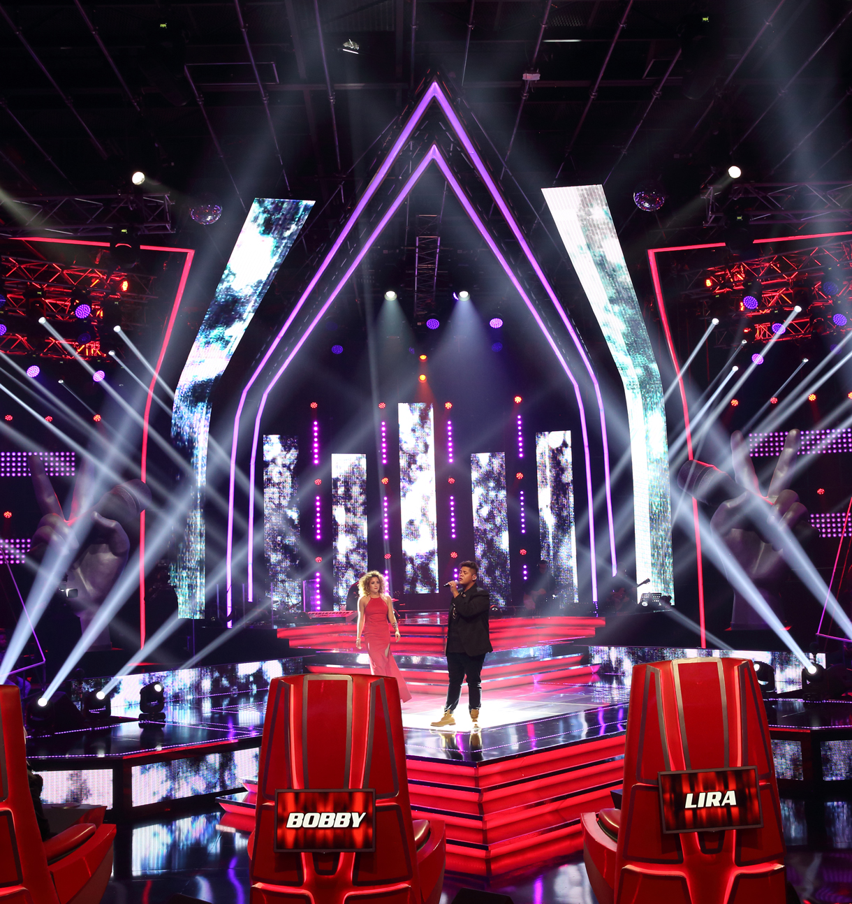 Portrait view of the Voice stage and red chairs beneath white beams.