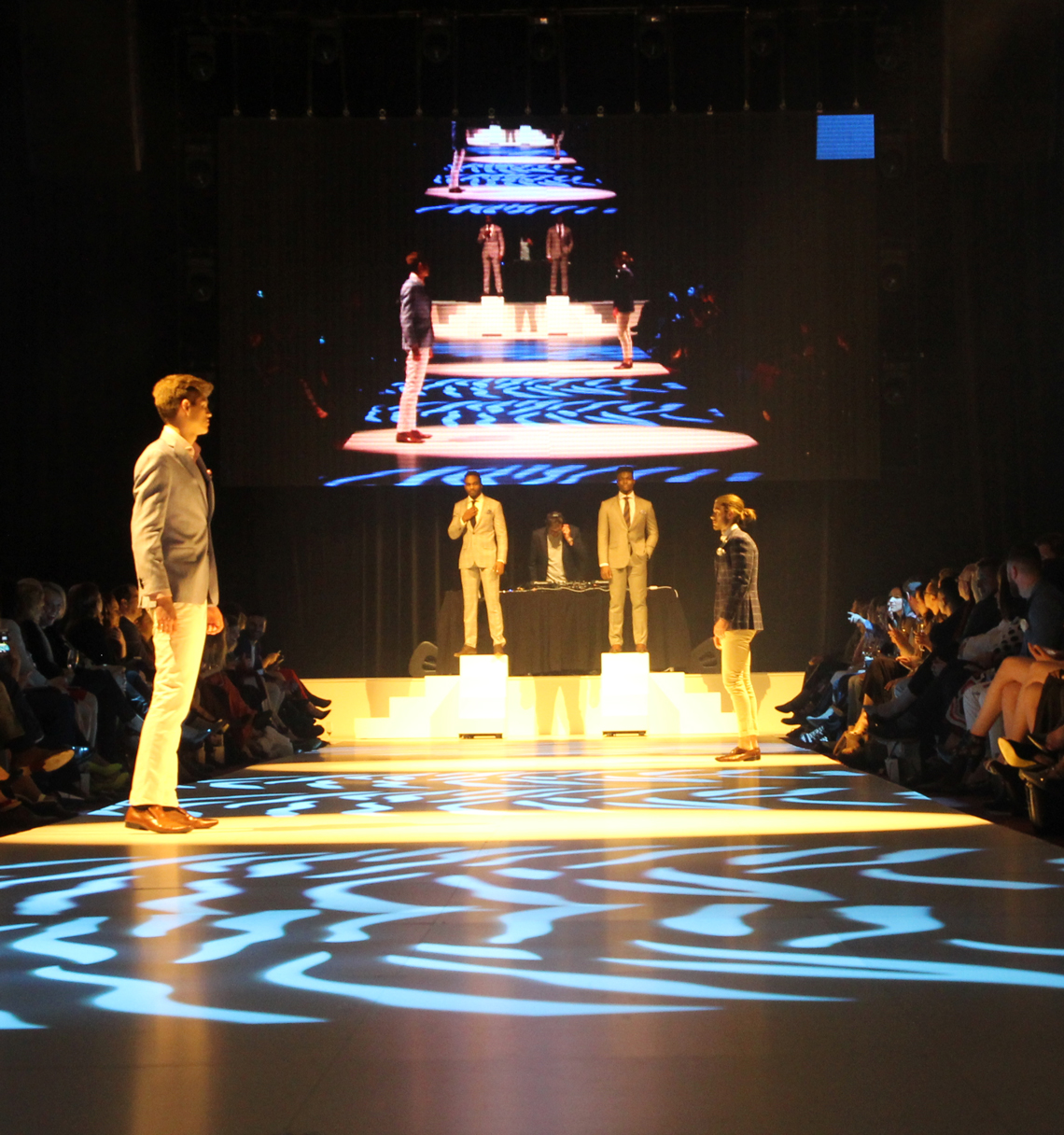 Models on a lit runway beside a tall tiered display at FashFest 2016.