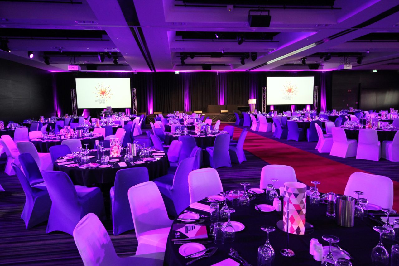 Wide view of a banquet at Adelaide Convention Centre with purple lighting and event screens.
