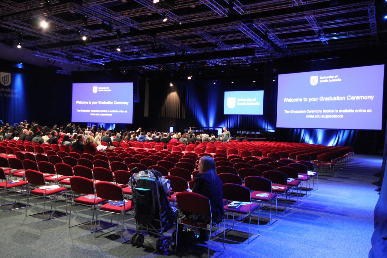 Rows of red conference chairs face three large presentation screens under blue lighting at Adelaide Convention Centre.