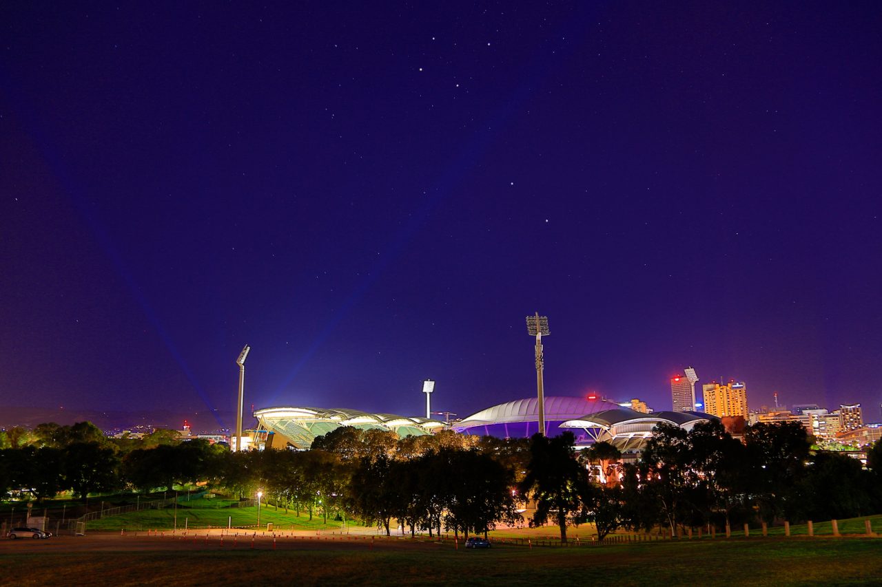 Adelaide Oval illuminated at night beyond trees and the river.