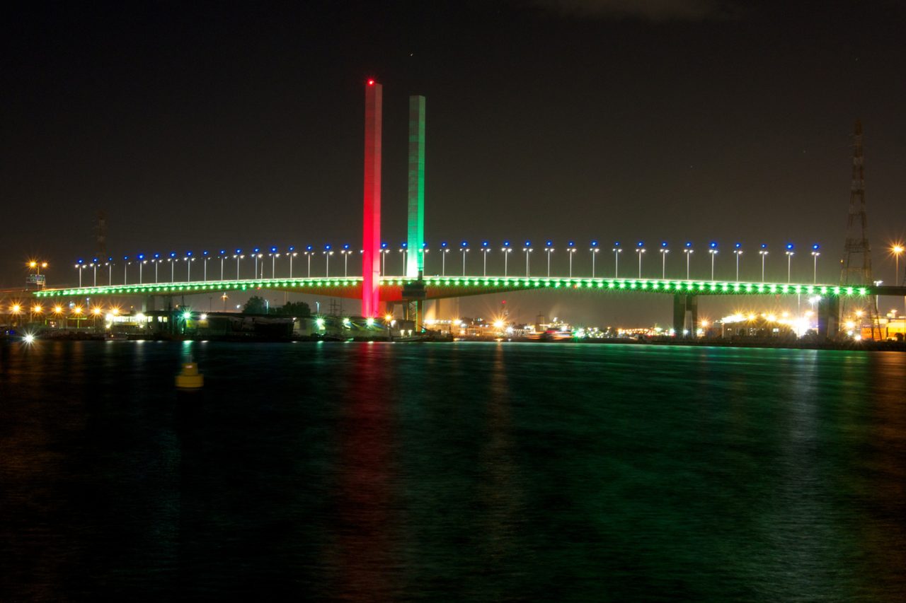 Red and green towers above the green-lit deck of Bolte Bridge at night.