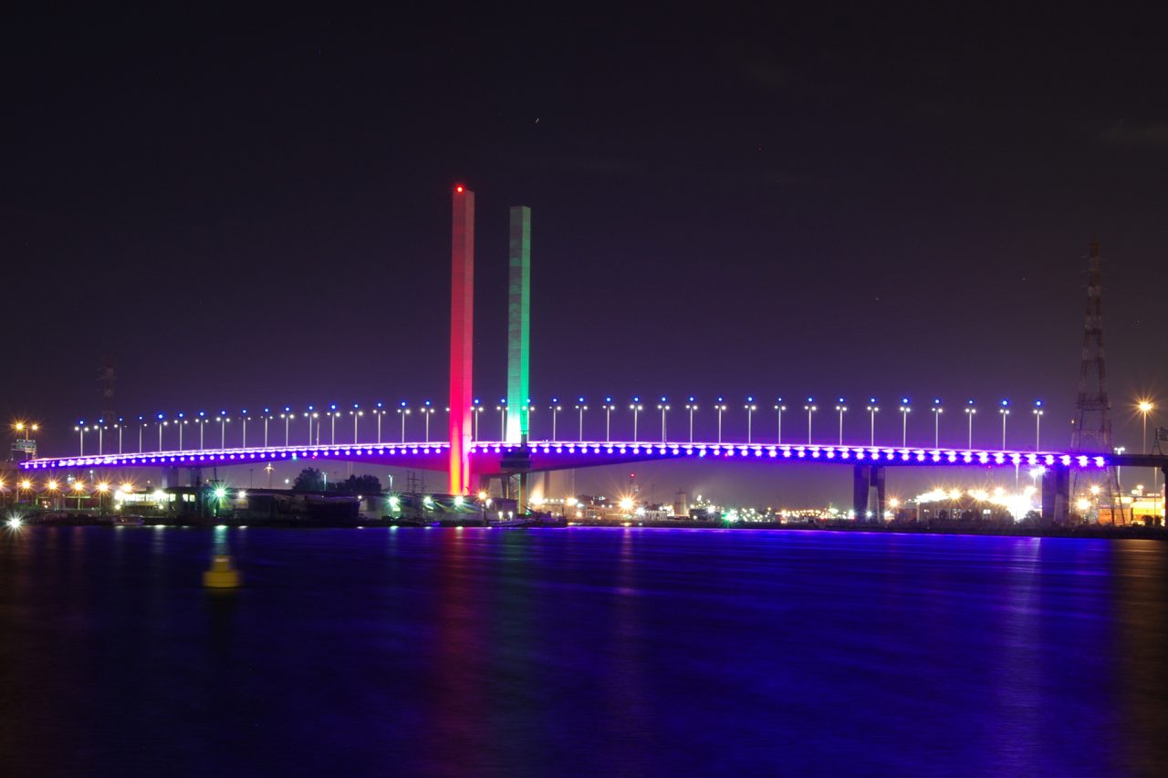 Bolte Bridge with red and green towers and a purple-lit deck reflected in the water.
