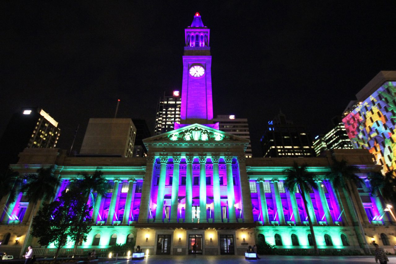 Brisbane City Hall clock tower and facade illuminated in purple and green.