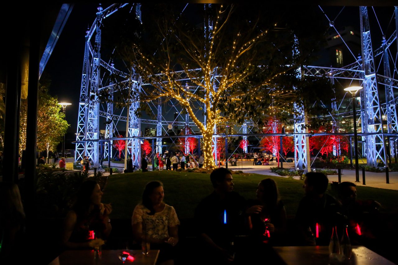 The gasometer framework at Gasworks Plaza lit blue and white behind trees at night.