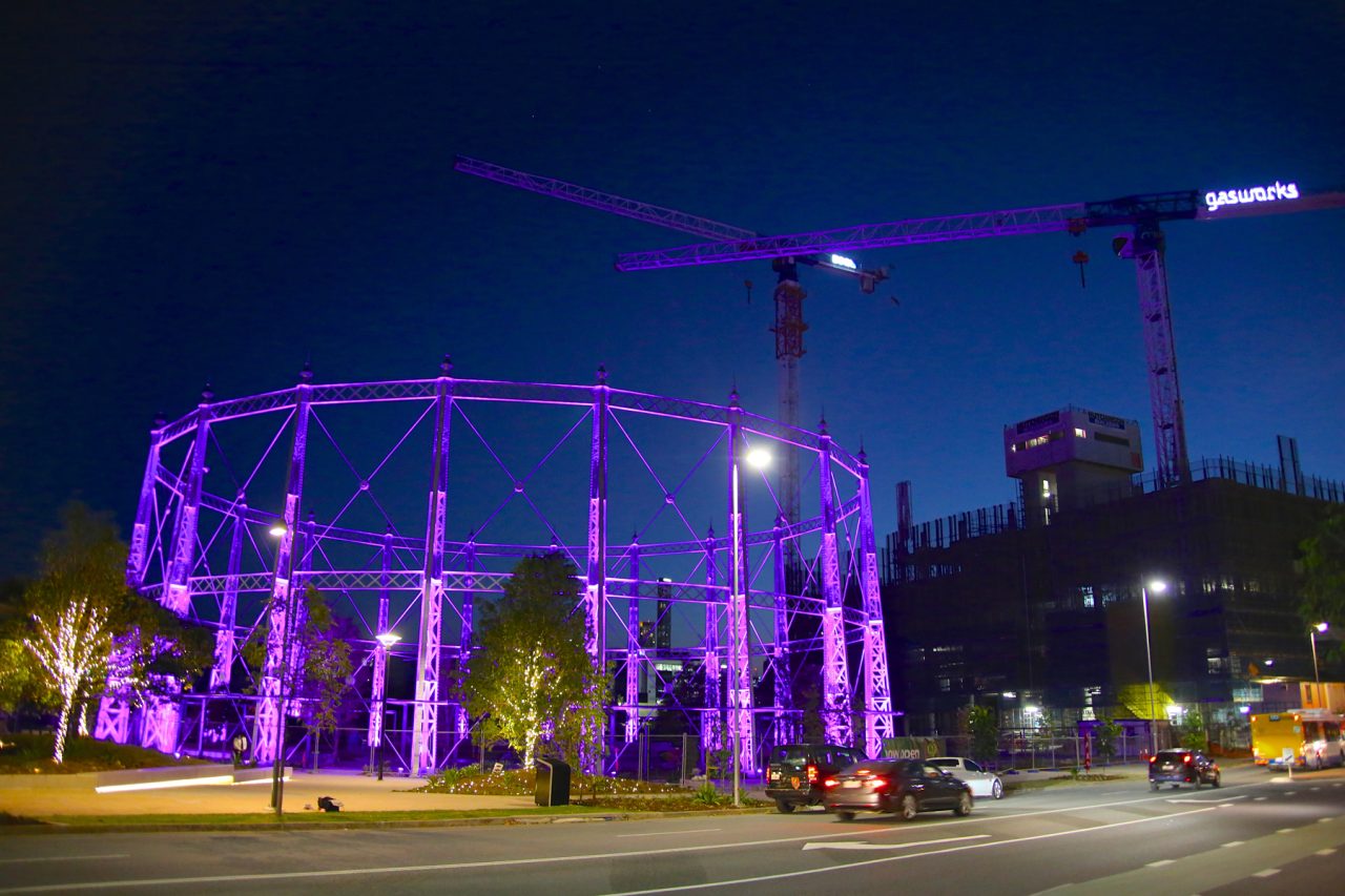 The circular gasometer at Gasworks Plaza illuminated purple across the road at night.