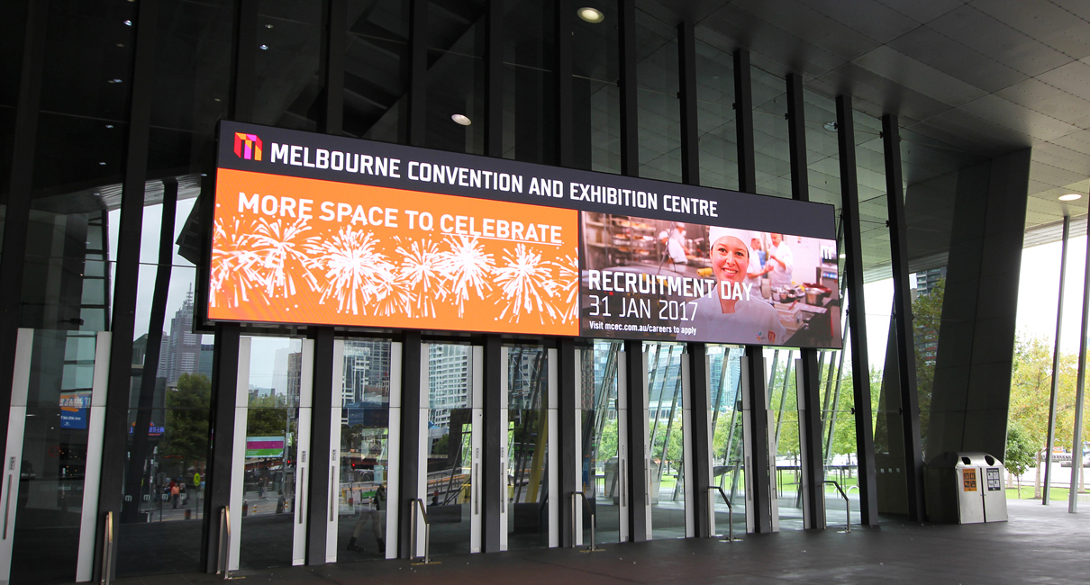 Orange event imagery across the outdoor display above the MCEC entrance.