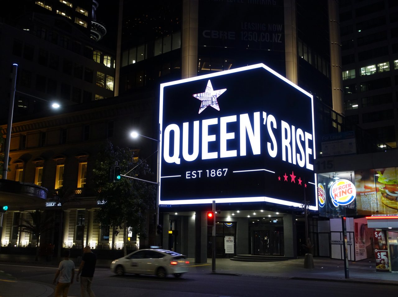 Queen's Rise branding illuminates the corner media facade in Auckland at night.