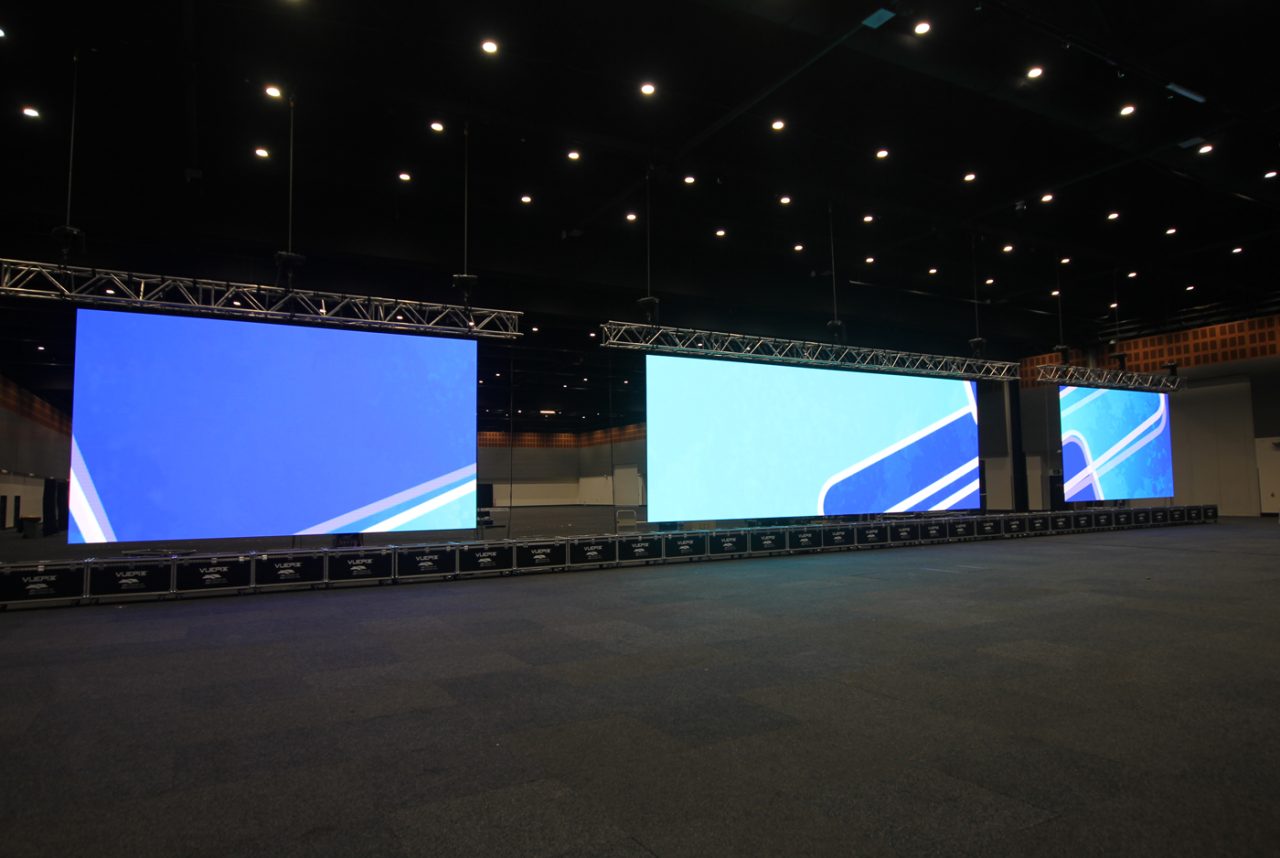 Three LED screens arranged across an empty convention hall.