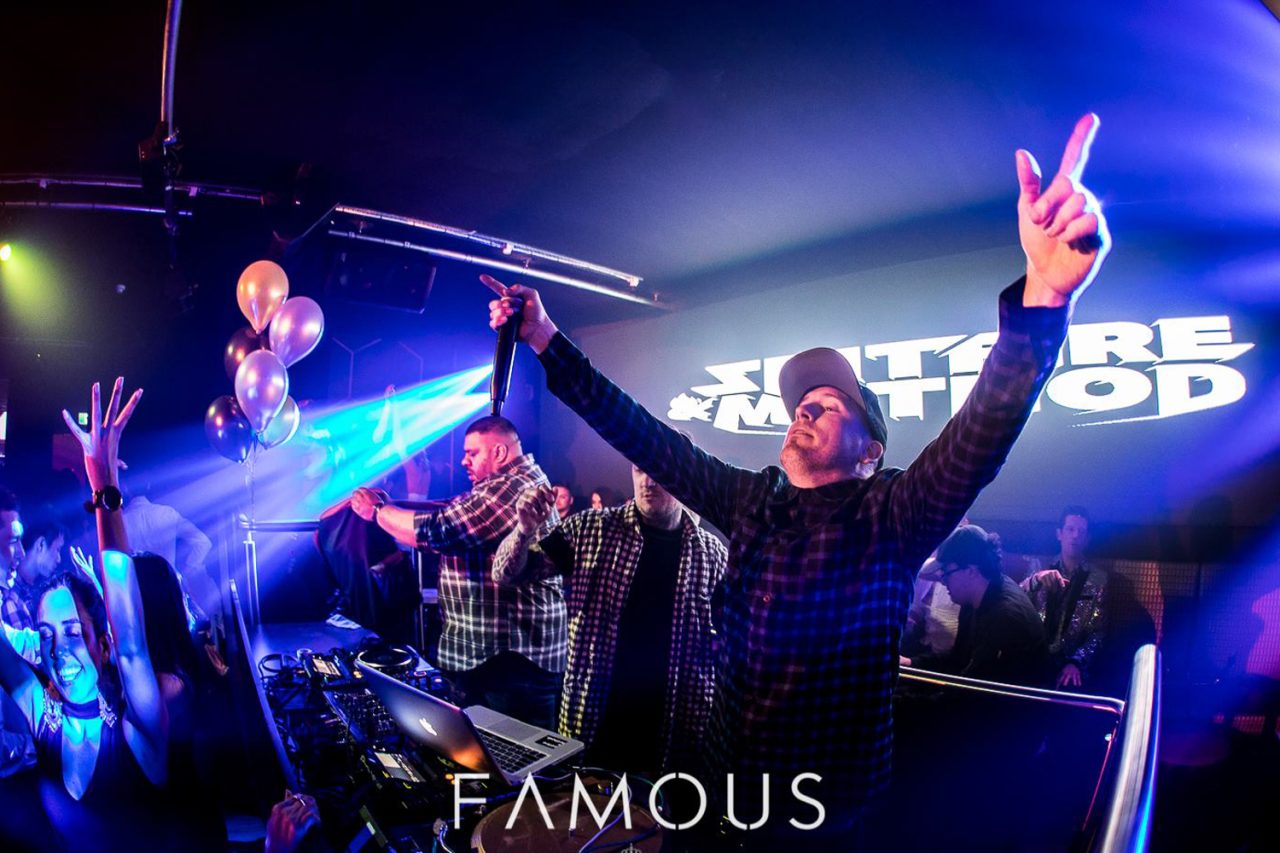 DJ raising both arms beneath blue beams at Famous Brisbane.