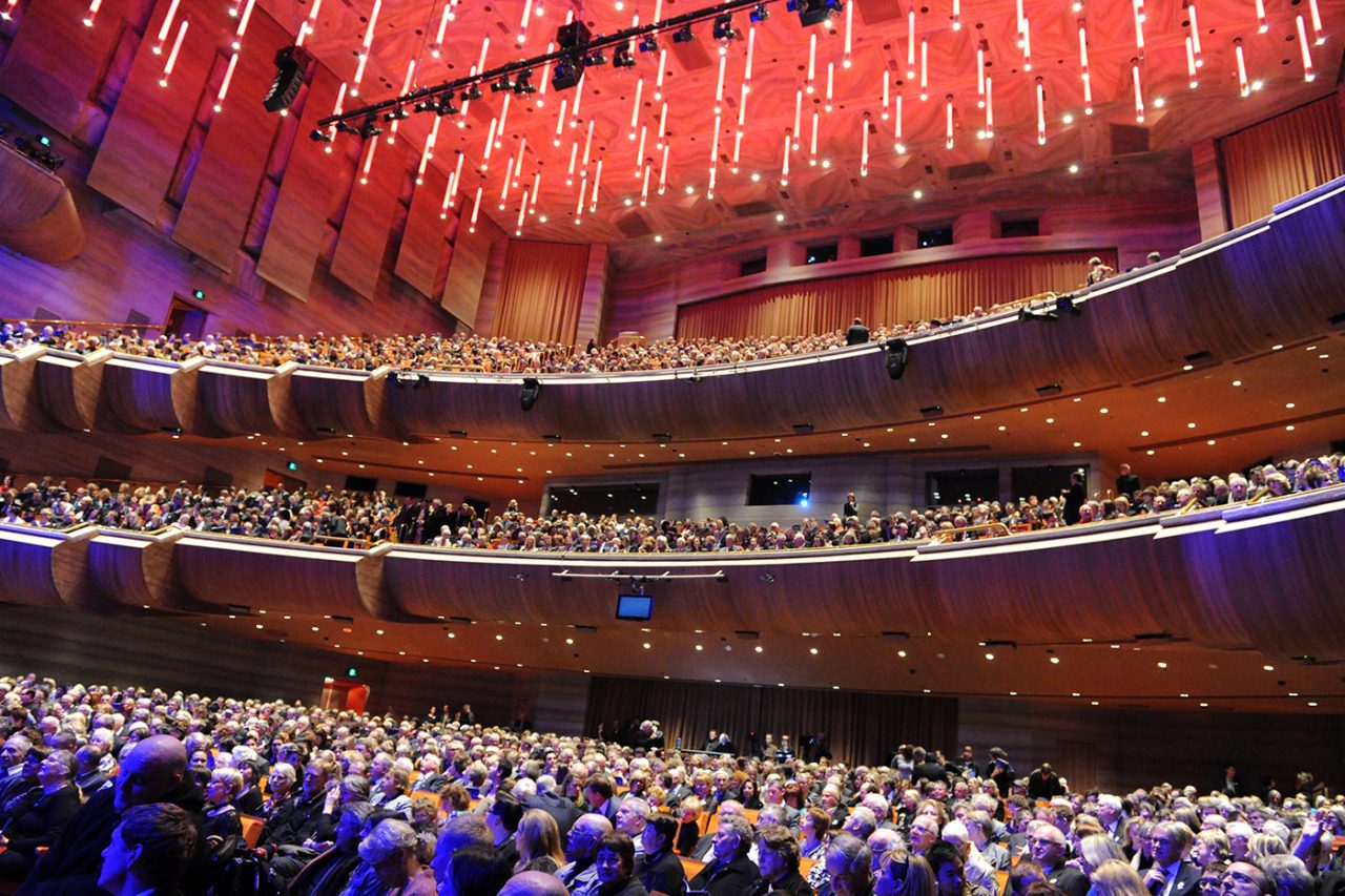 Audience fills Hamer Hall beneath warm ceiling lighting and purple balcony accents.