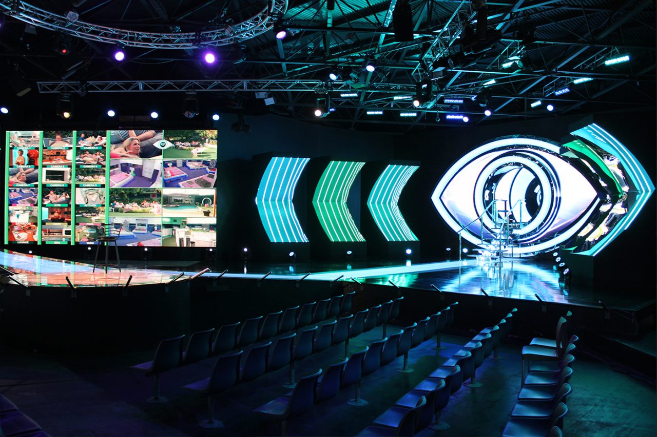 Wide view of the Big Brother studio, audience seating and eye-shaped screen graphics.