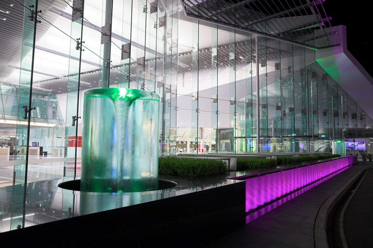 Cylindrical water feature glowing green beside a purple-lit channel at Canberra Airport.