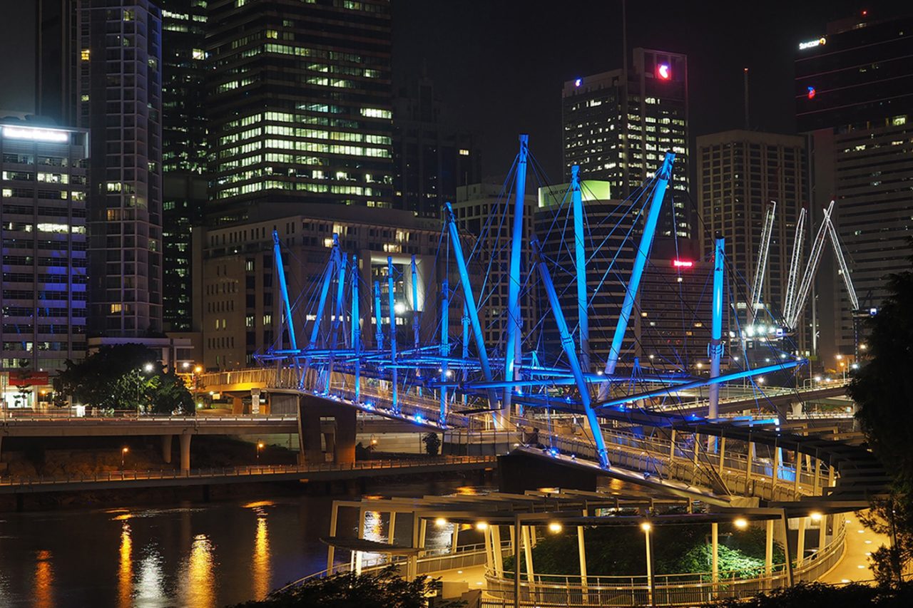 Blue-lit masts on Kurilpa Bridge reflected in the river at night.