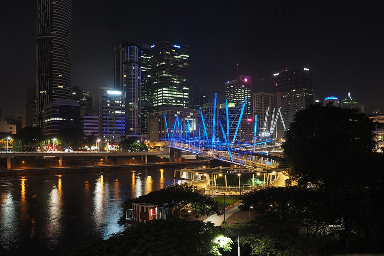 Wide view of blue-lit Kurilpa Bridge and Brisbane's night skyline.