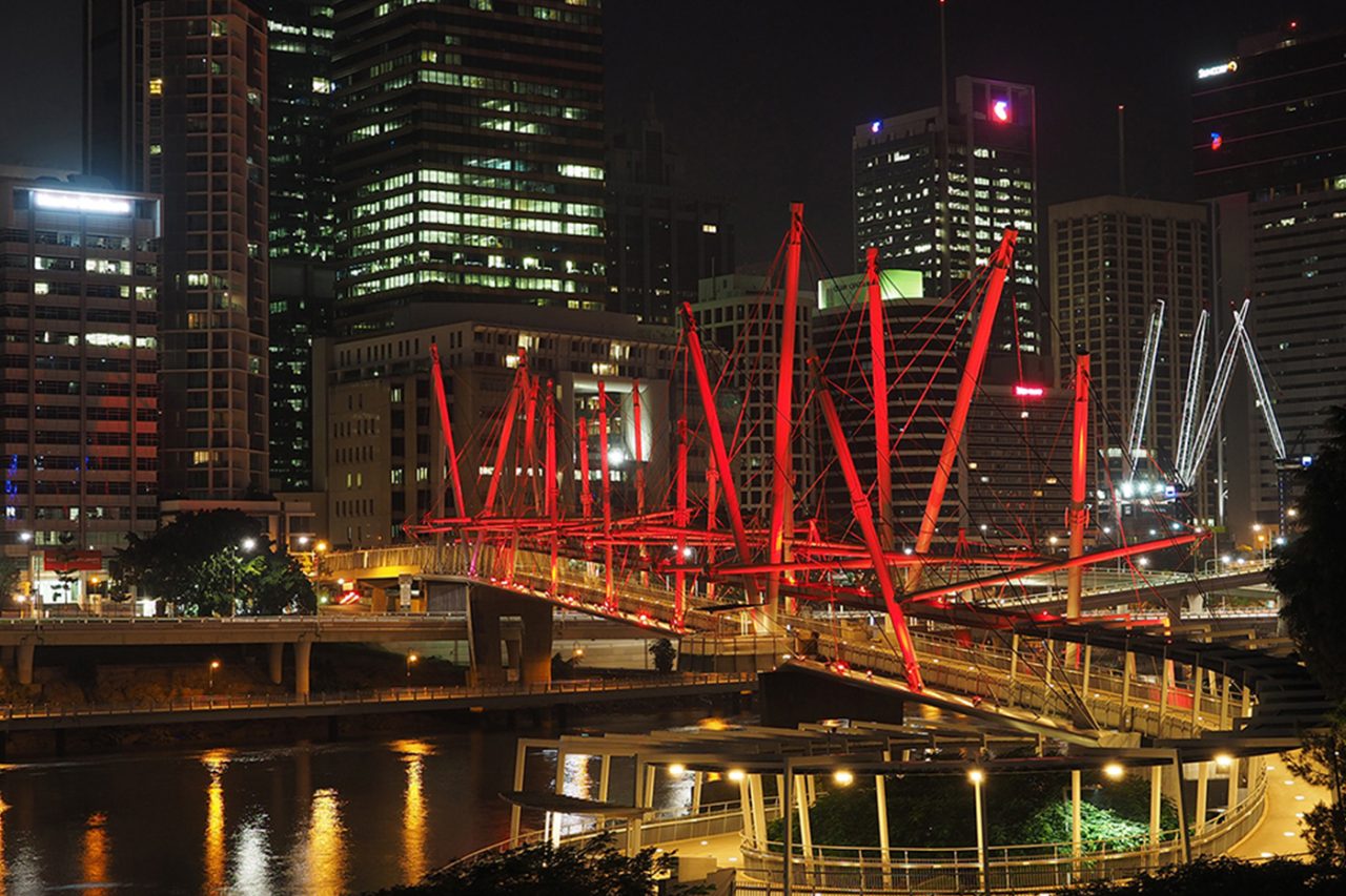 Kurilpa Bridge lit red beside the Brisbane city skyline at night.