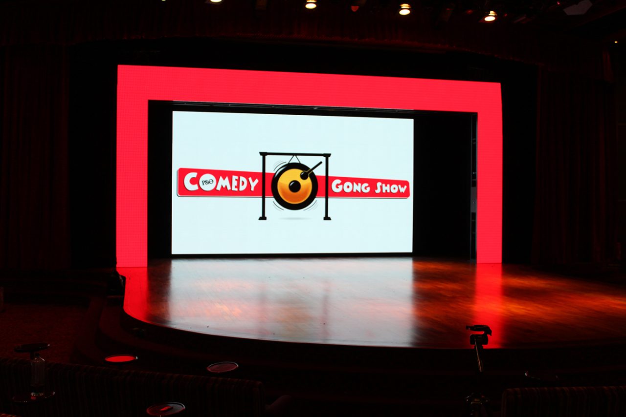 Comedy branding on a stage screen surrounded by a red digital frame aboard Pacific Dawn.
