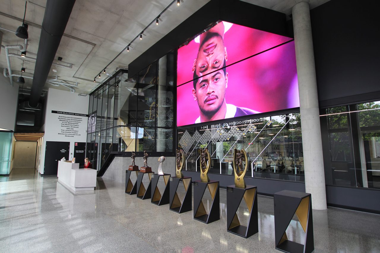 Portrait footage on the foyer screen above the Brisbane Broncos trophy display.