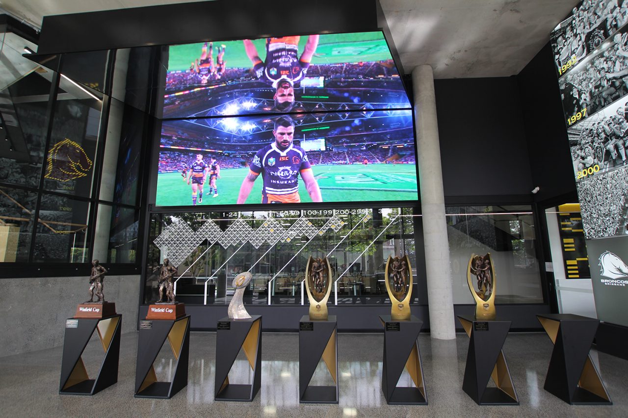 Rugby league footage on the large screen behind the Broncos trophy display.