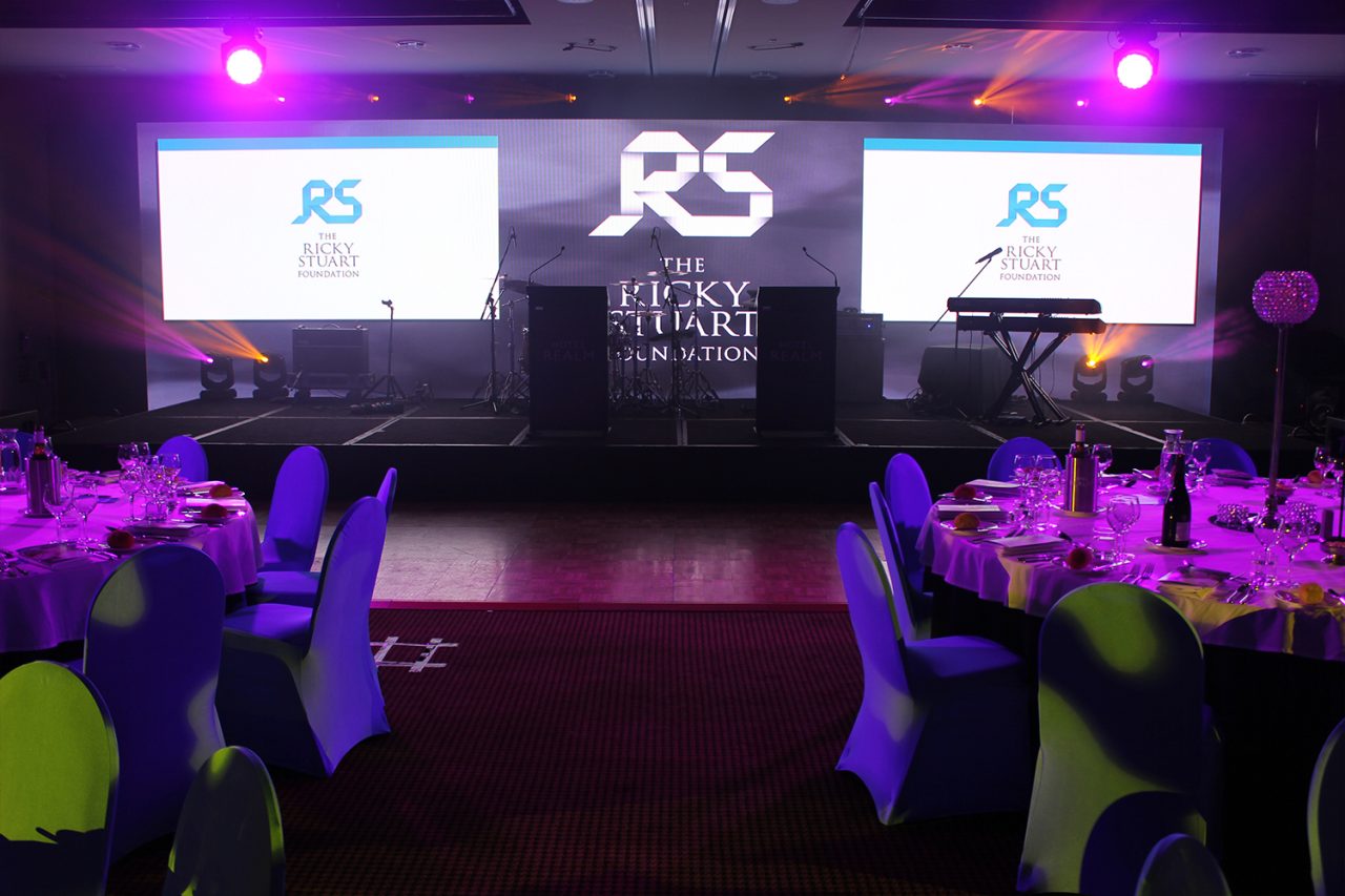 Wide view of the gala stage and branded screens between banquet tables.