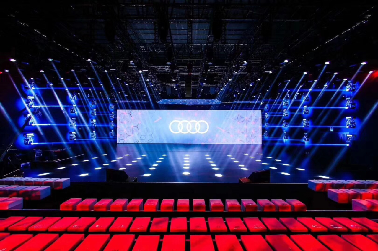 Blue-lit Audi launch stage with a central screen and rows of red seats.