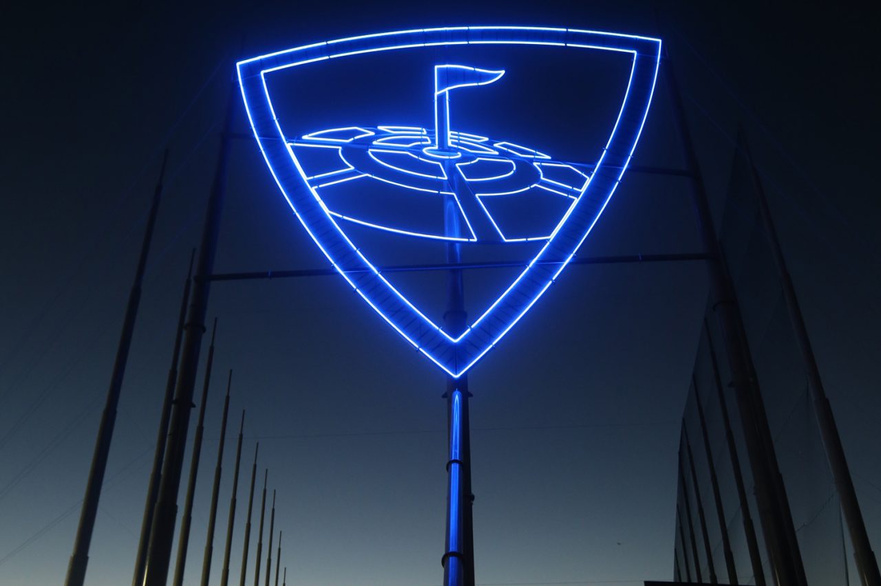 Blue illuminated Topgolf shield sign above the range at night.