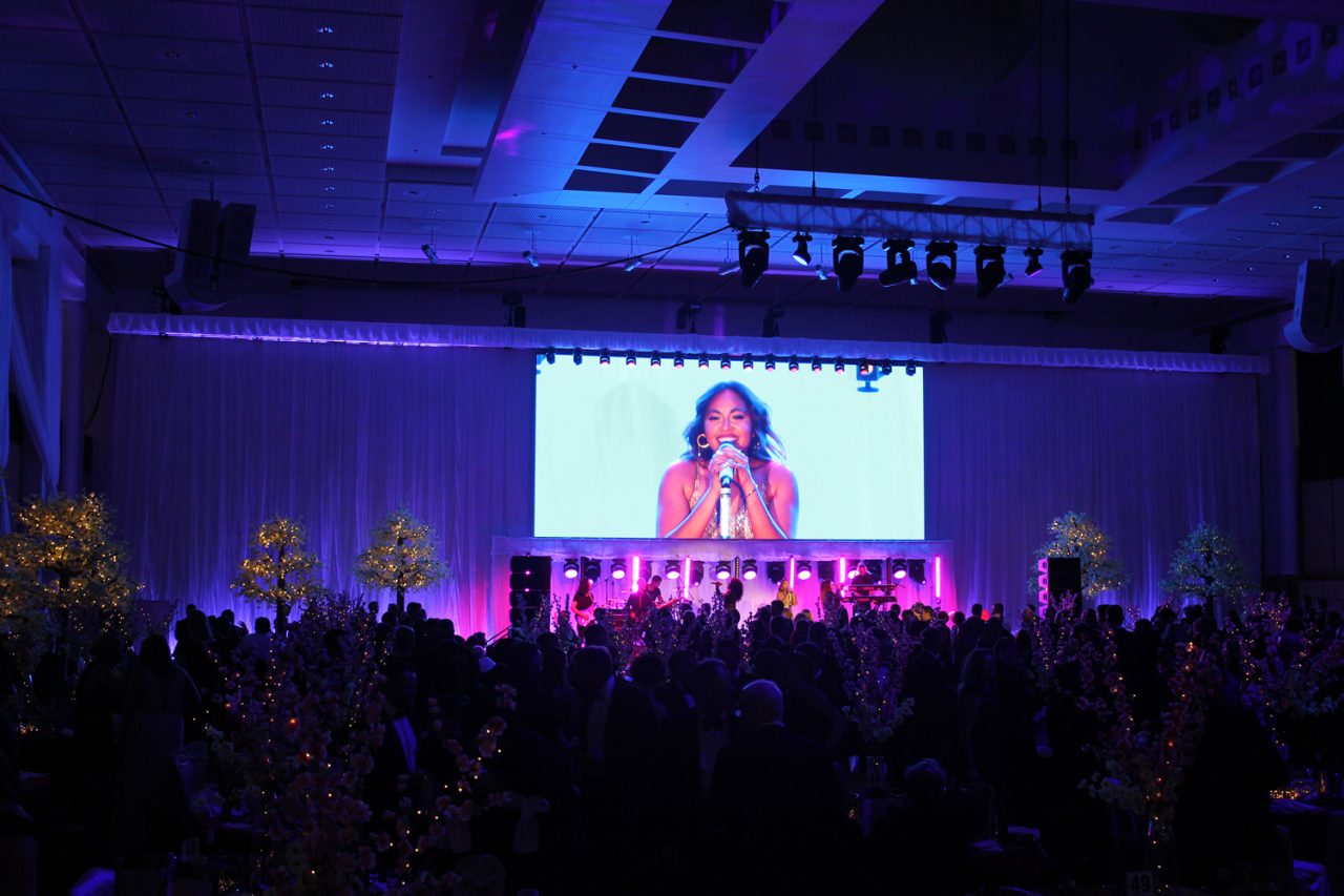 A live speaker shown on the central screen in the blue-lit Mid Winter Ball venue.