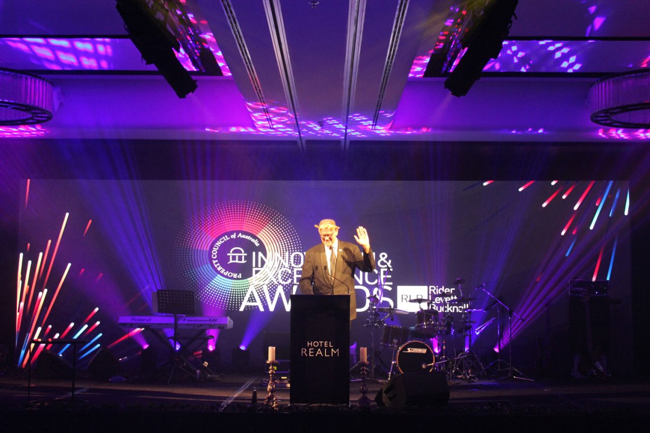 Presenter at a lectern in front of Property Council ACT Awards screen graphics.