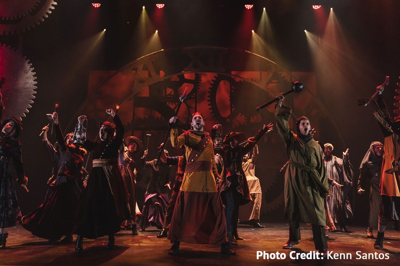 Wicked cast on a warm-lit stage with a mechanical backdrop at HOTA.