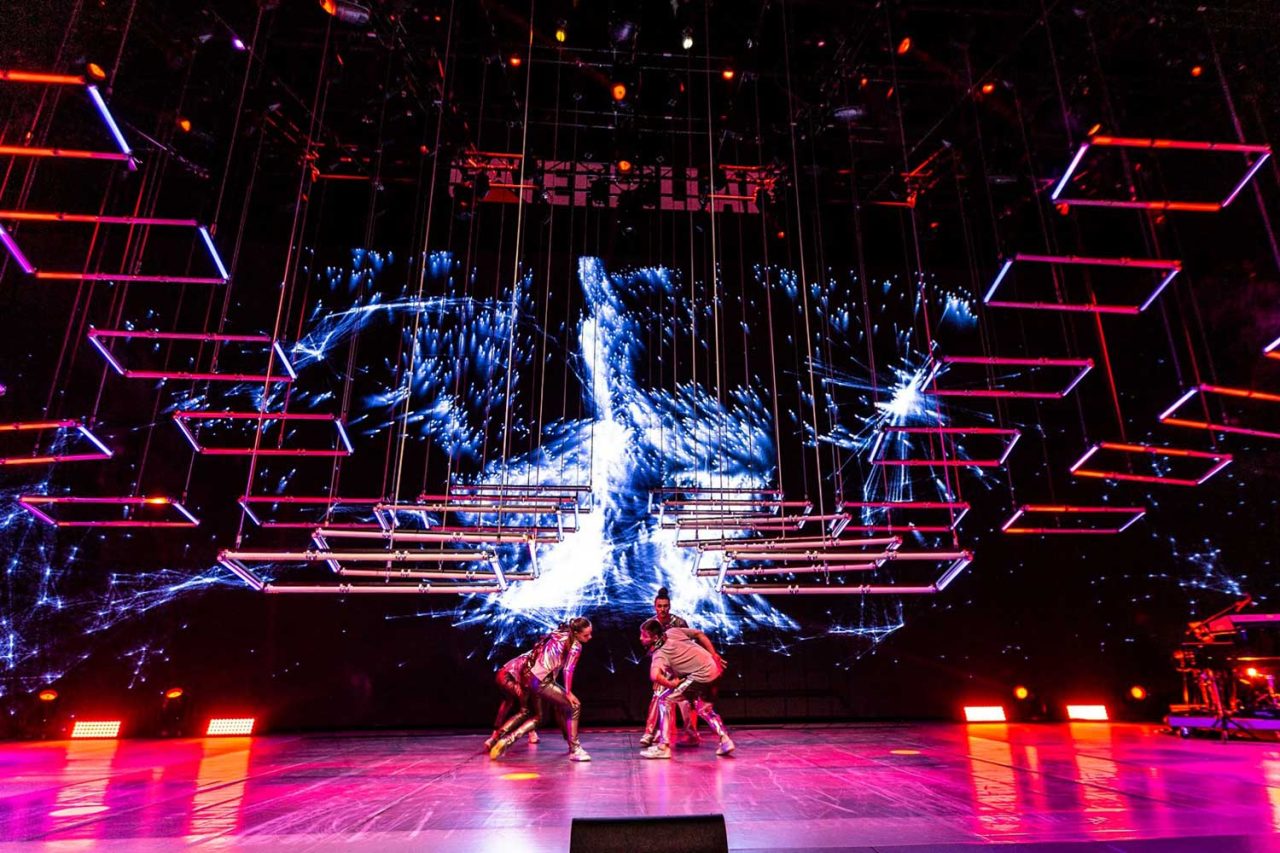 Performer onstage beneath geometric red and blue tube lighting.