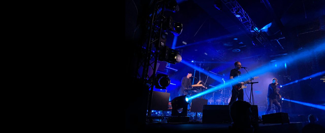 Band performing on a dark stage crossed by blue beams.