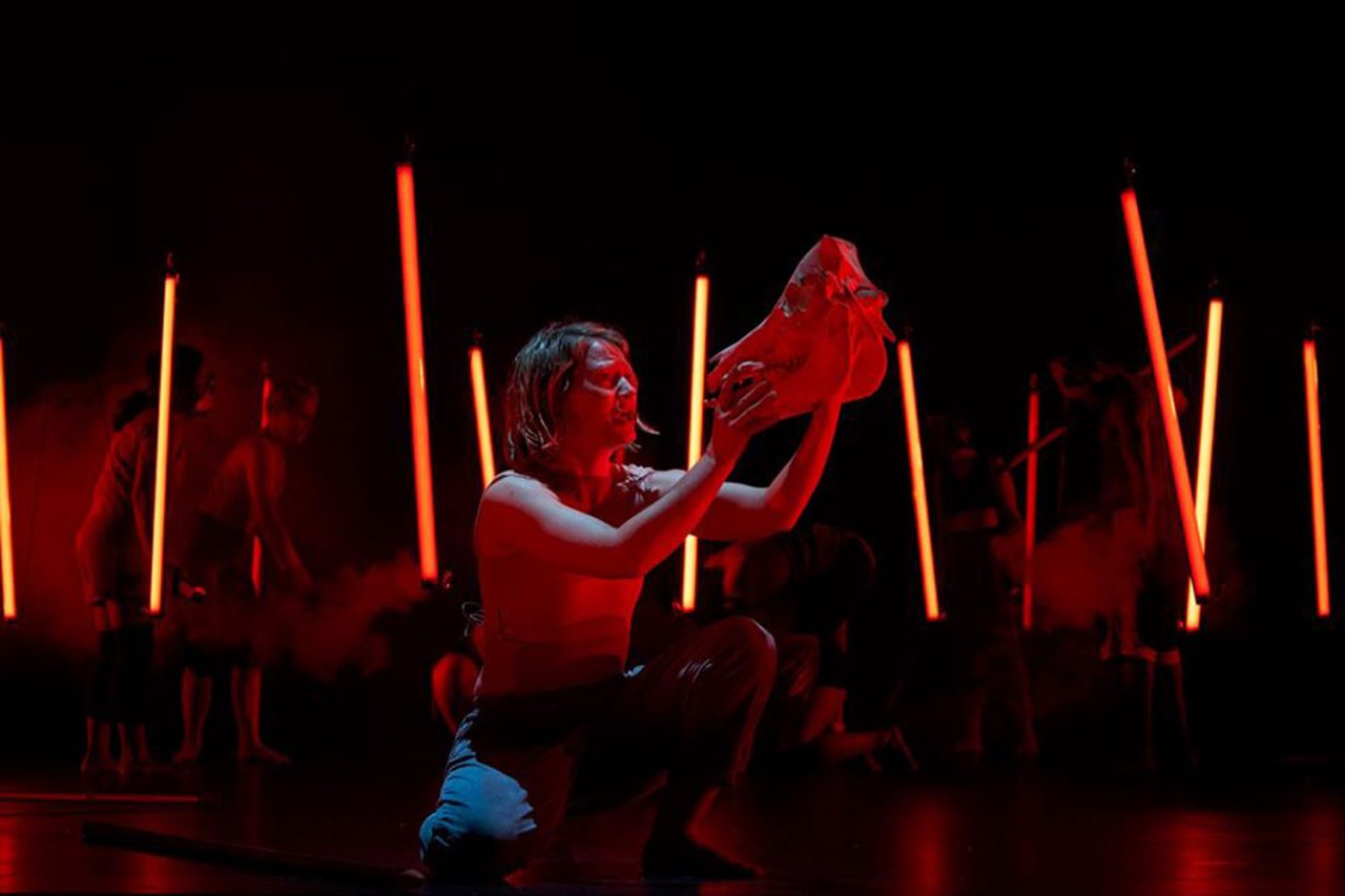Red light tubes surround a performer holding a prop in Lord of the Flies.