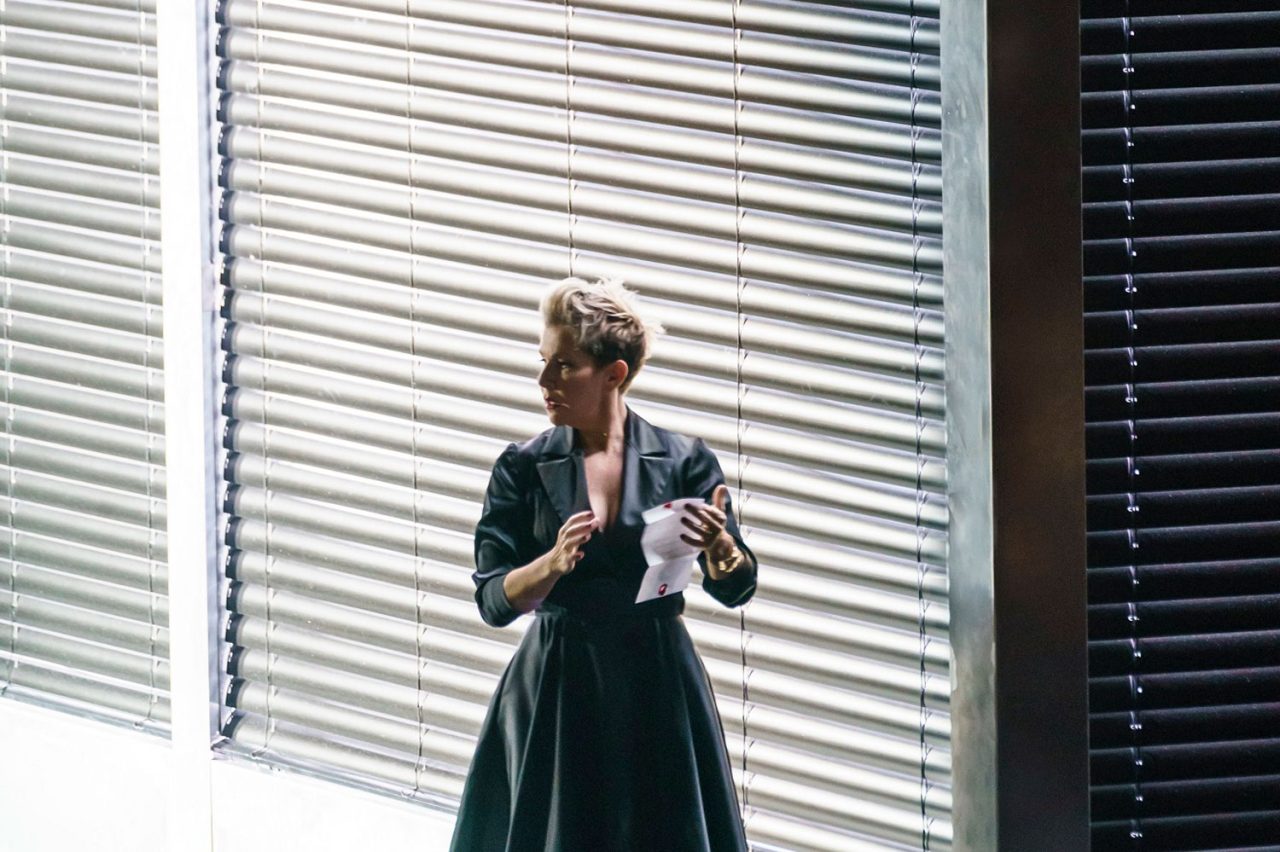 Performer standing beside large louvred window panels onstage.