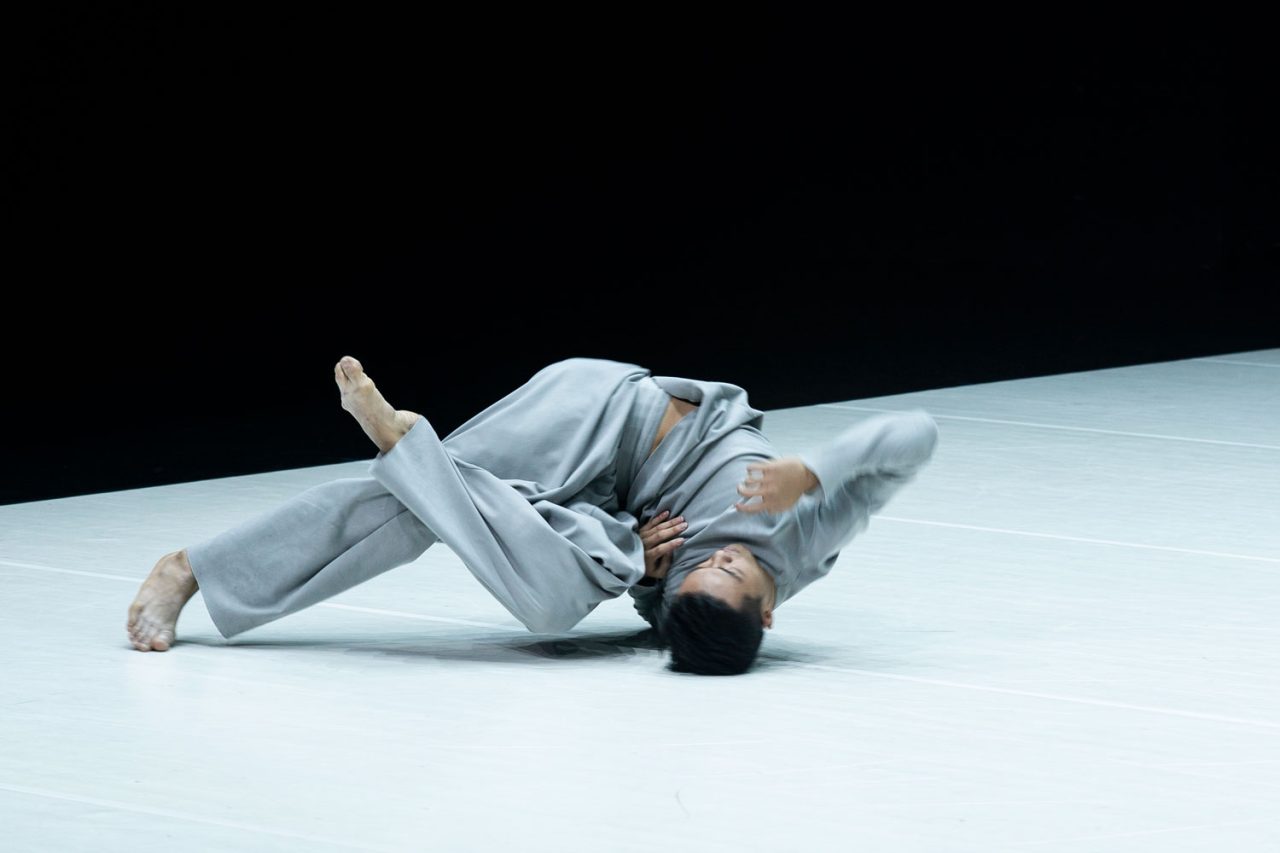 Dancer in a pale costume curled on a brightly lit white stage.