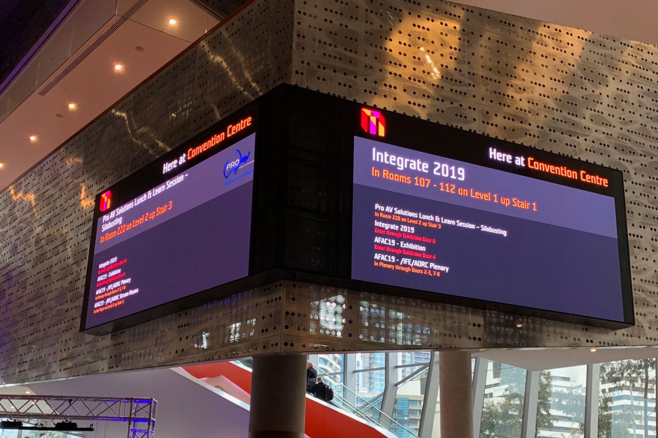 Paired digital event information screens above an escalator in the MCEC foyer.