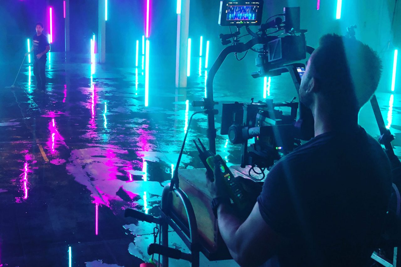 Camera operator among cyan and magenta light tubes on a reflective floor.