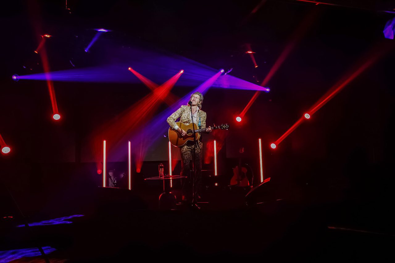 Guitarist onstage beneath red beams and vertical light tubes.