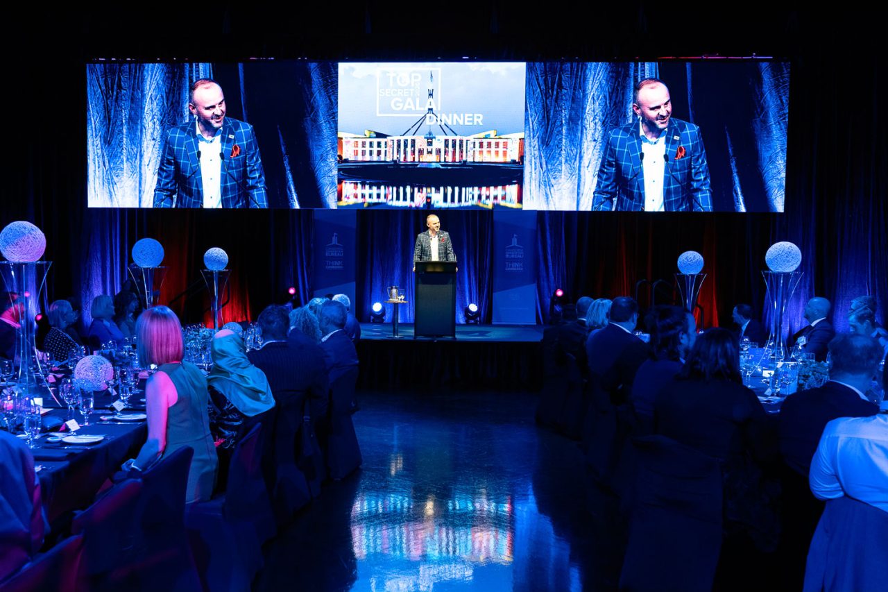 Speaker at a lectern beneath live camera screens at the Top Secret gala.