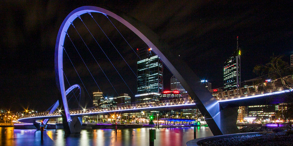 Illuminated arch bridge and waterfront buildings reflected in the water at night.