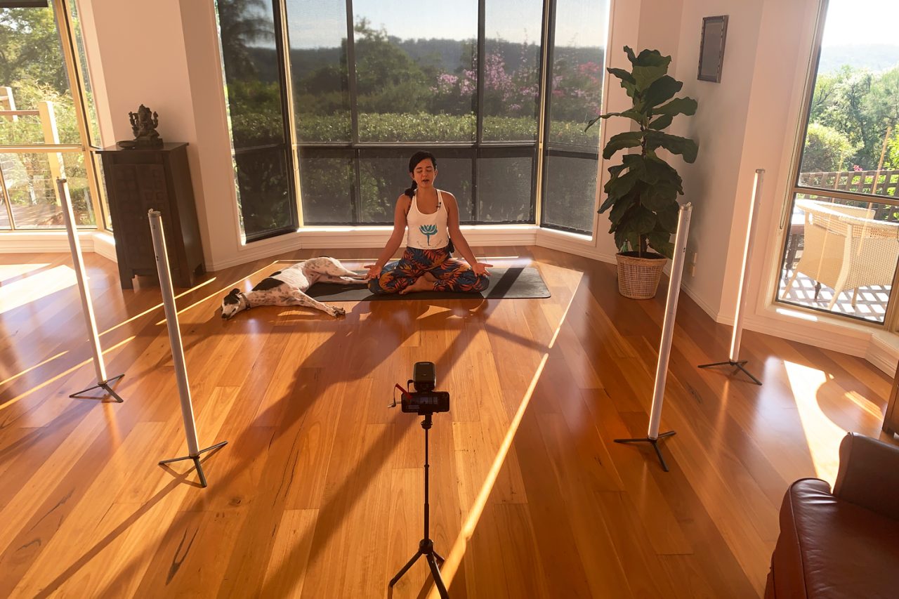 Instructor seated on a mat near windows with a camera and tube lights for streaming.