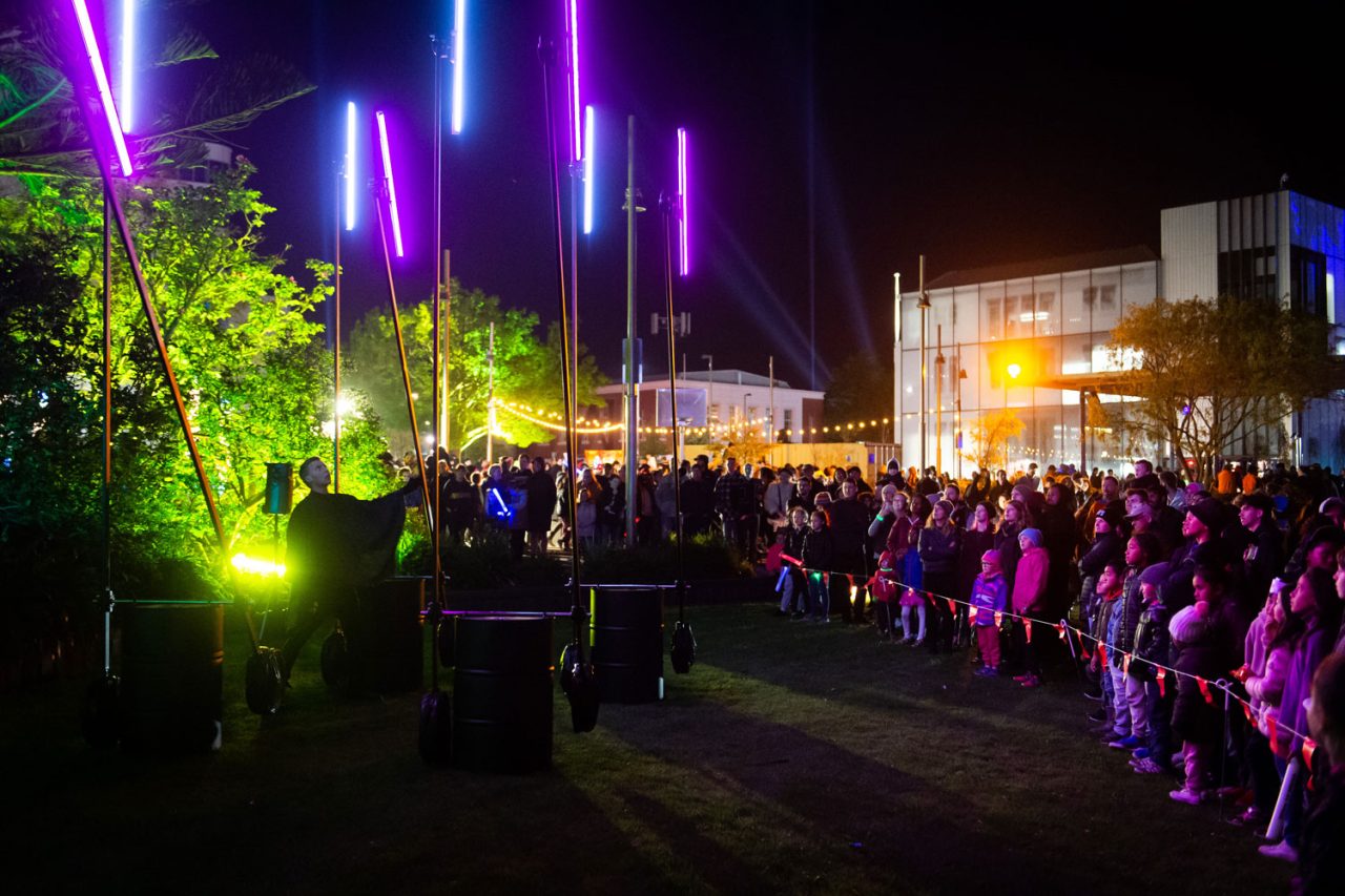 Audience watches an outdoor performance beneath suspended colourful light tubes.