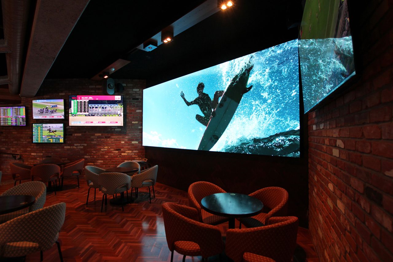 Large screen at 1st on Wharf Tavern showing a surfer above the lounge seating.
