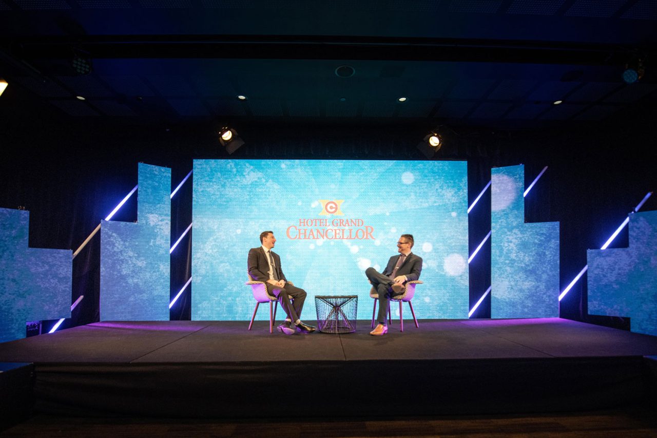 Two people seated on a Scene Change virtual-studio stage in front of a large blue display.