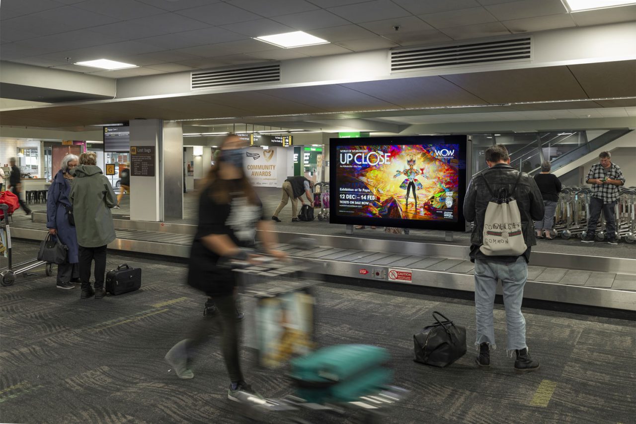 Advertising screen beside the baggage-carousel area at Wellington Airport, with passengers passing in front.