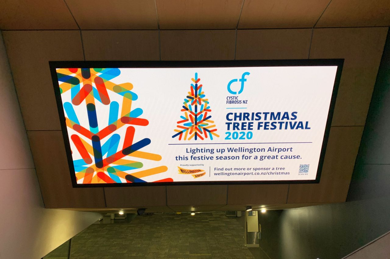 Wellington Airport advertising screen displaying a Christmas Tree Festival promotion.