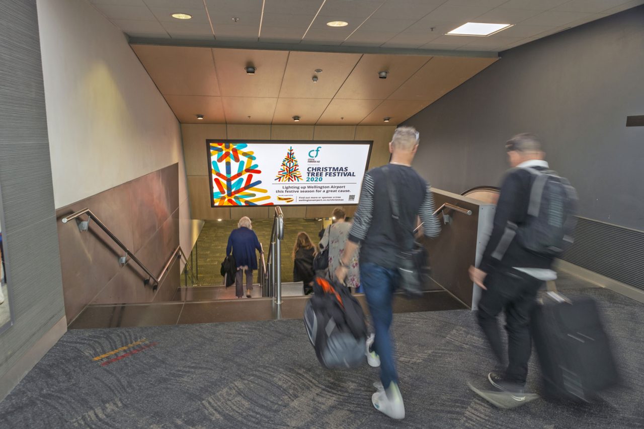 Passengers beneath an overhead advertising display at Wellington Airport.