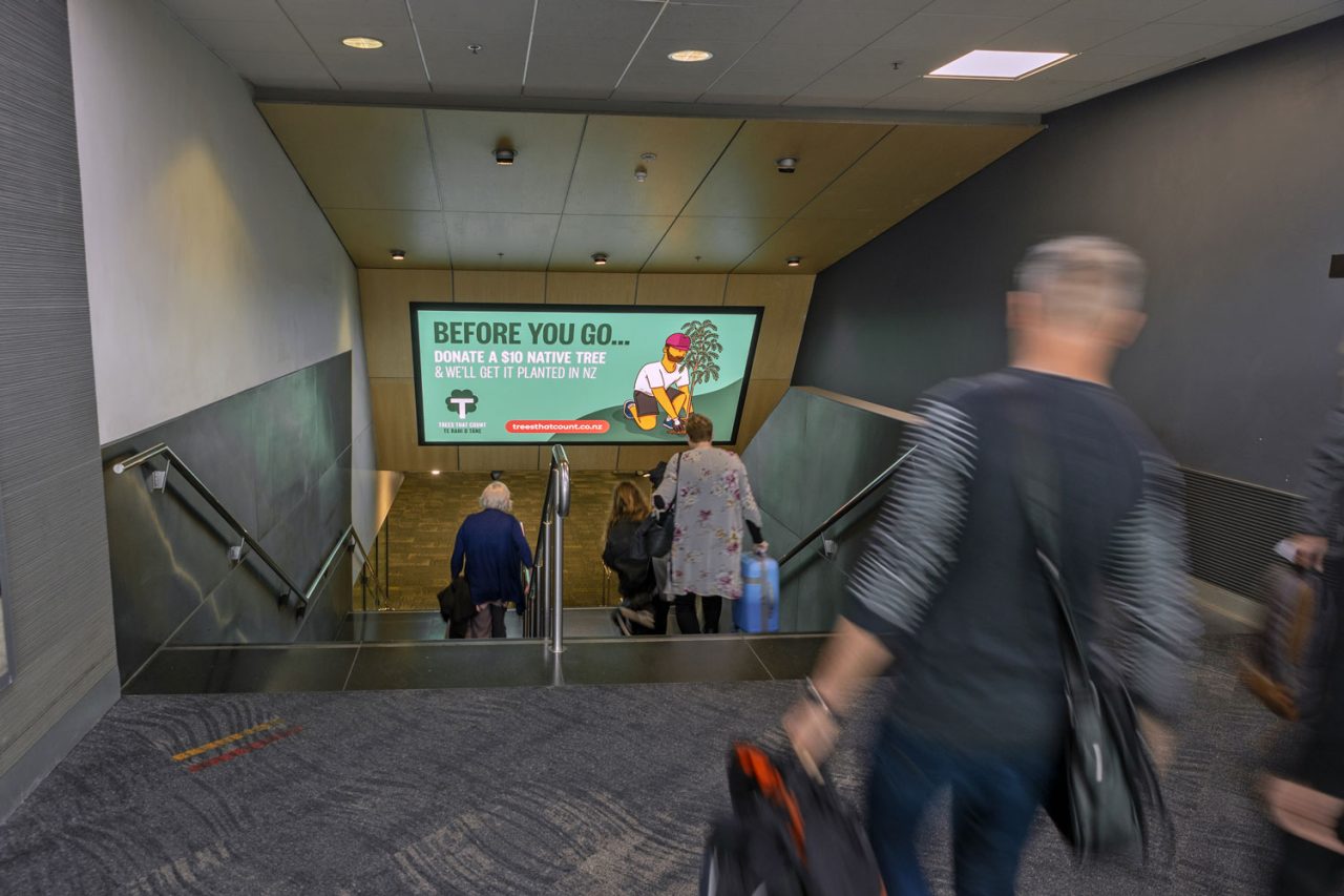 Passengers descending stairs beneath an advertising screen at Wellington Airport.