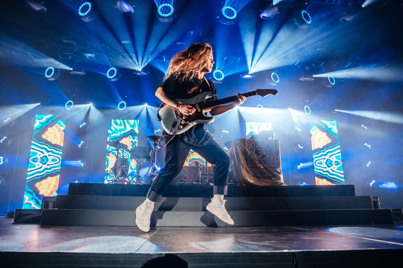 Tash Sultana performing with a guitar beneath blue stage beams and colourful displays.