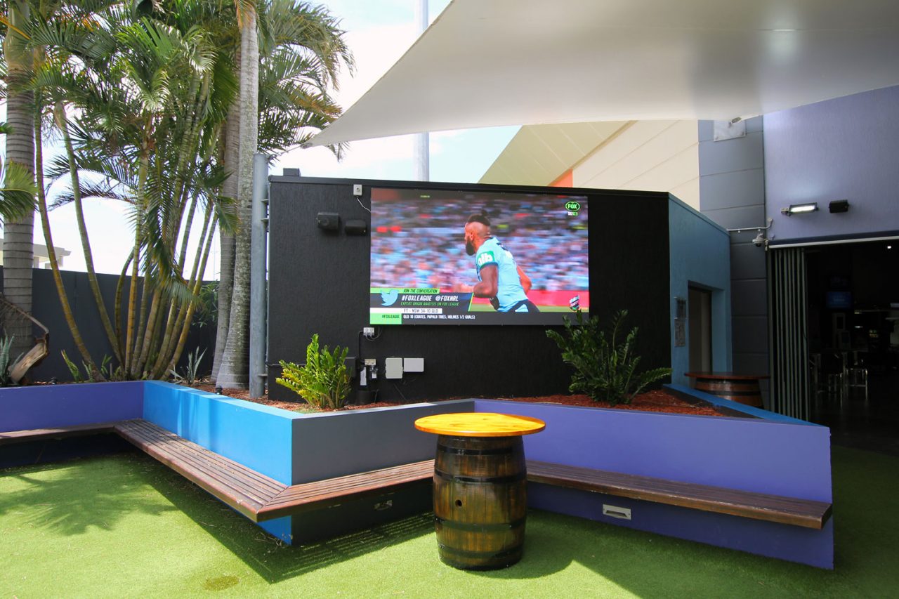 Large outdoor screen framed by blue benches and palm trees at Redcliffe Leagues Club.