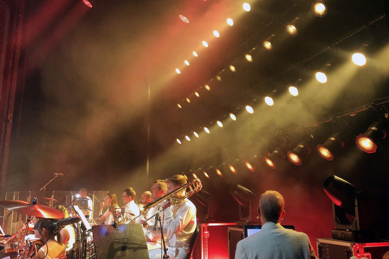 Side-stage view of performers beneath warm beams at Bohemian Symphony.